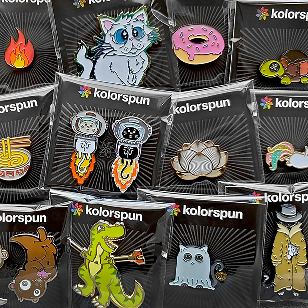 Enamel pins on Kolorspun display cards showing a fire emoji, blue-eyed cat, pink donut, and dinosaurs.