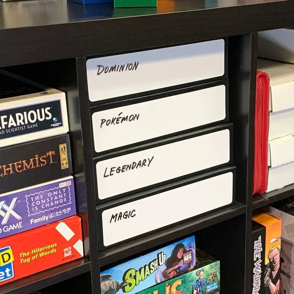 Black 3-row card game box with white accents, designed by THE MIGHTY HOBBY SHOP, on display on a shelf.