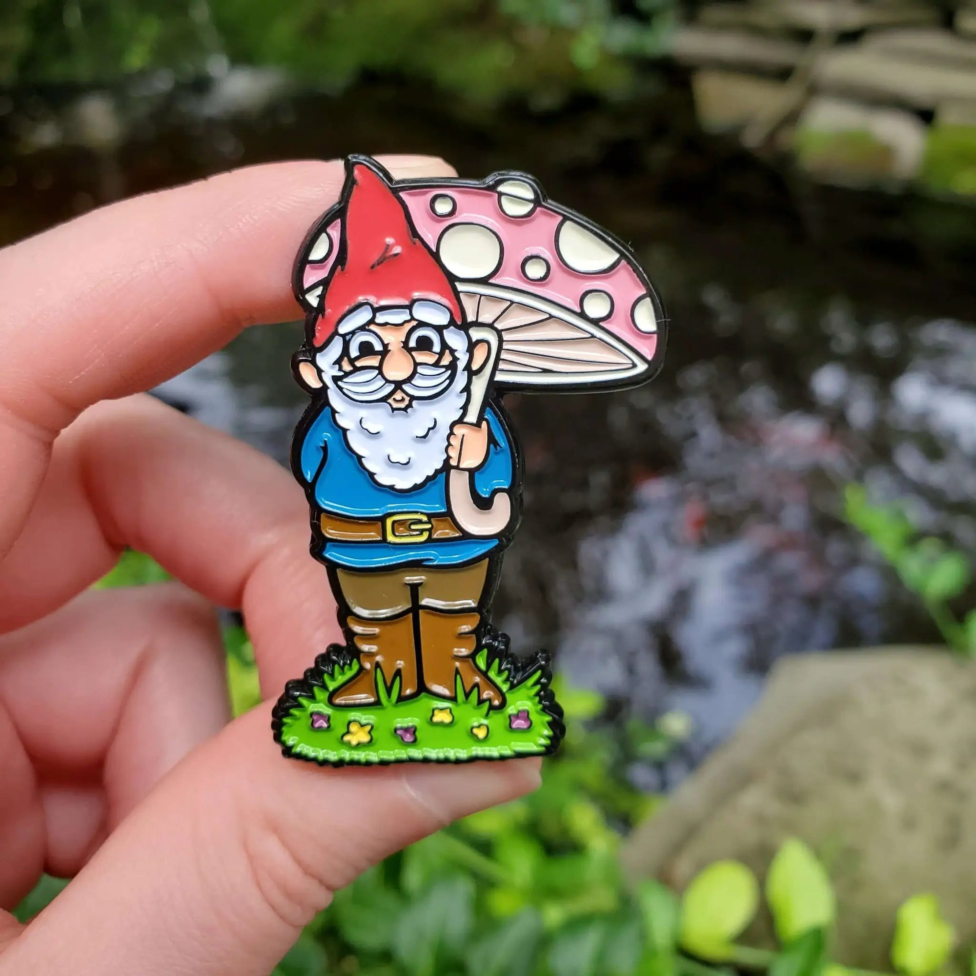 Kolorspun garden gnome enamel pin featuring a whimsical gnome figure sitting near a pond, perfect for outdoor-themed accessories.