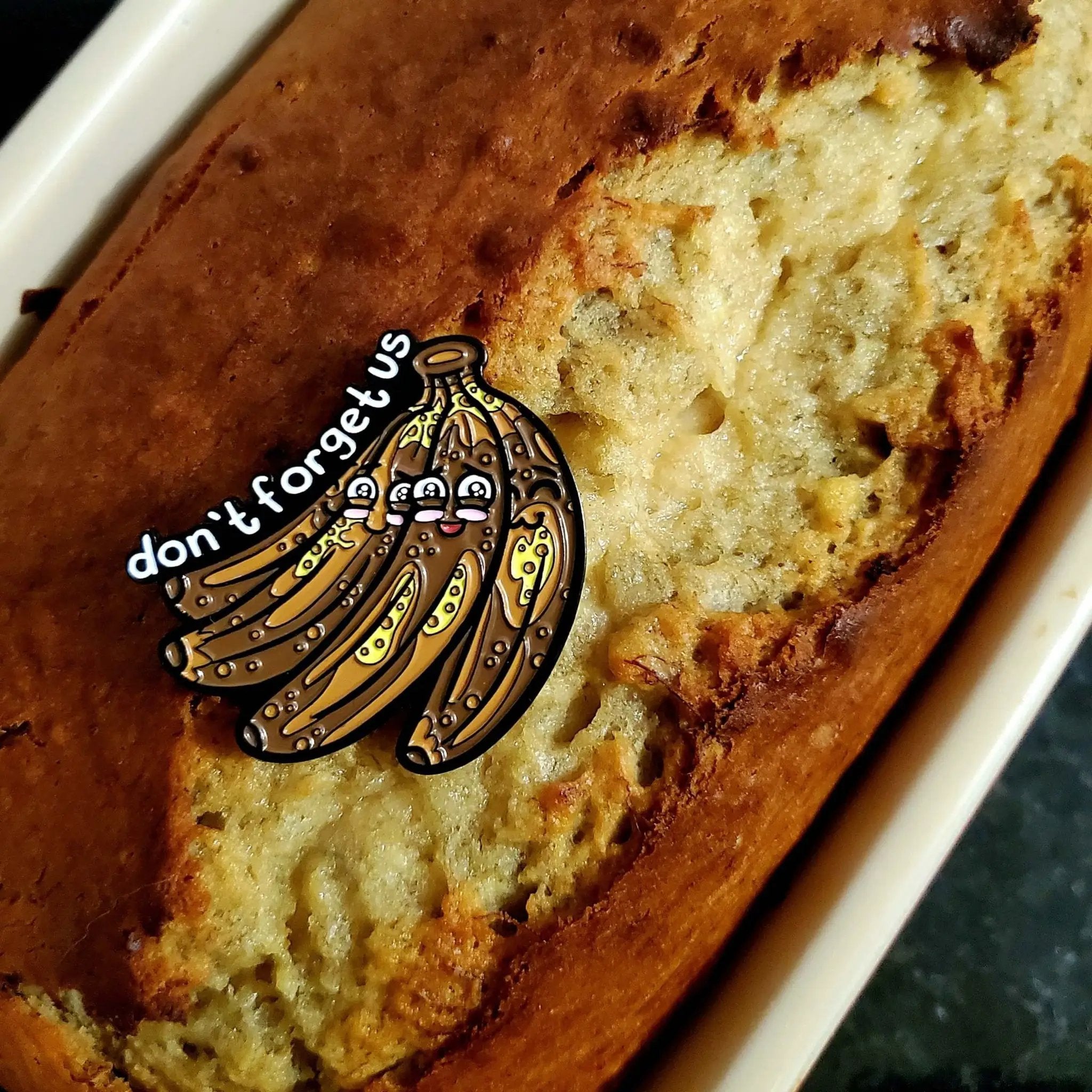 Kolorspun "Don't Forget Us" Bananas enamel pin on banana bread background, showcasing colorful pin design and playful style.
